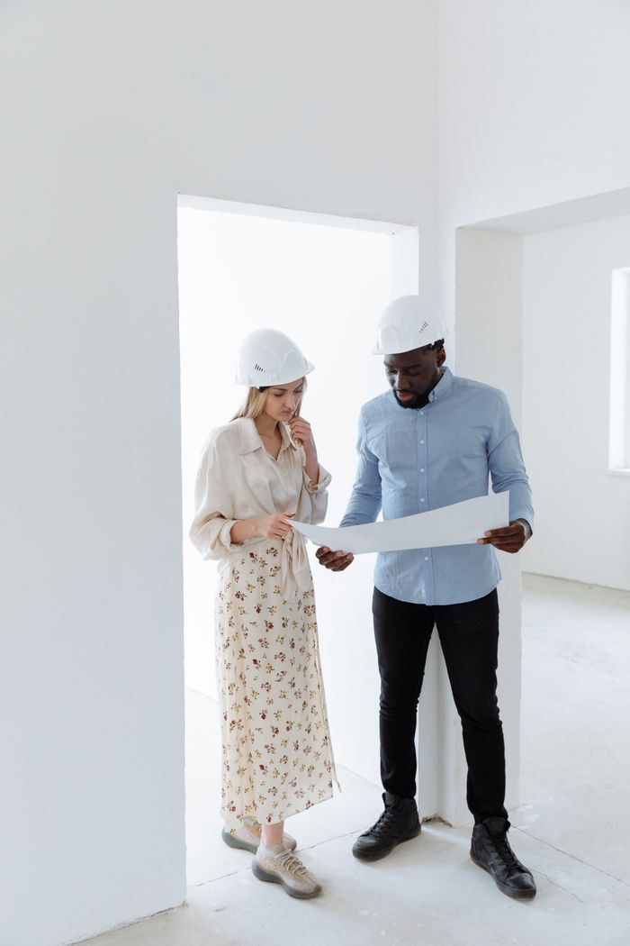 Two architects in hard hats examine a blueprint at a construction site indoors.